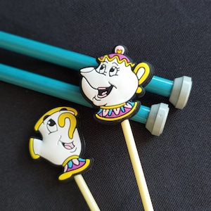 Może przedstawiać: Dwie niebieskie druty do robótek na drutach z białymi końcówkami i żółtymi uchwytami. Uchwyty są ozdobione głowami postaci z filmu Disneya Piękna i Bestia. Jeden uchwyt przedstawia Panią Czajnik, czajnik, a drugi uchwyt przedstawia Chip, filiżankę do herbaty.