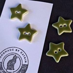 May include: Four olive green star-shaped buttons with two holes each. Two buttons are attached to a white rectangular card featuring a black circular stamp with a cat silhouette and the text "HANDMADE BY CATKIN MADE WITH LOVE".