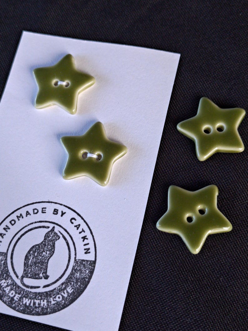 May include: Four olive green star-shaped buttons with two holes each. Two buttons are attached to a white rectangular card featuring a black circular stamp with a cat silhouette and the text "HANDMADE BY CATKIN MADE WITH LOVE".