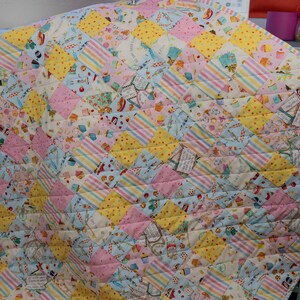 May include: A colorful quilt with a patchwork design featuring a variety of fabrics with a cupcake theme. The quilt is made up of squares of different colors and patterns, including pink, yellow, blue, and white. The quilt is quilted with a diamond pattern.