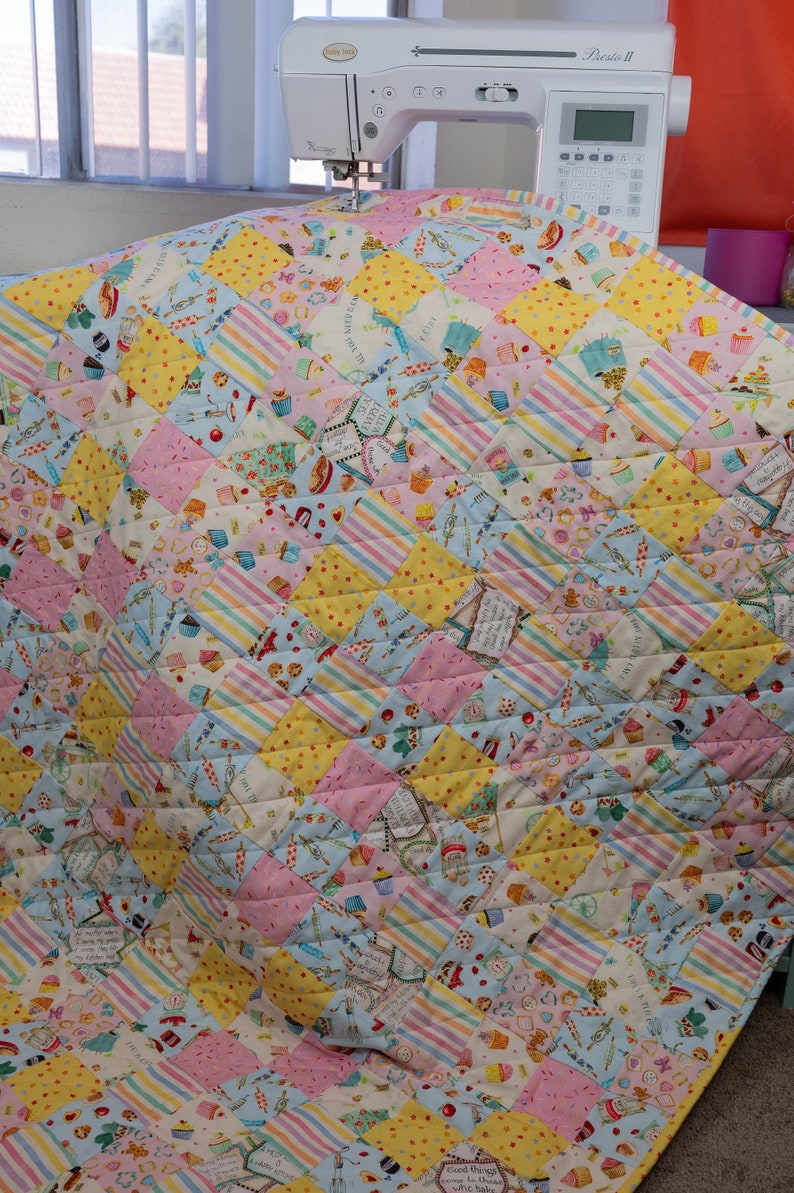 May include: A colorful quilt with a patchwork design featuring a variety of fabrics with a cupcake theme. The quilt is made up of squares of different colors and patterns, including pink, yellow, blue, and white. The quilt is quilted with a diamond pattern.