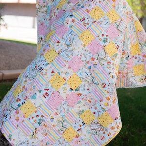 May include: A patchwork quilt with a light blue, yellow, and pink color scheme. The quilt is made of various fabrics with different patterns, including stripes, polka dots, and floral designs. The quilt is hanging on a tree branch.