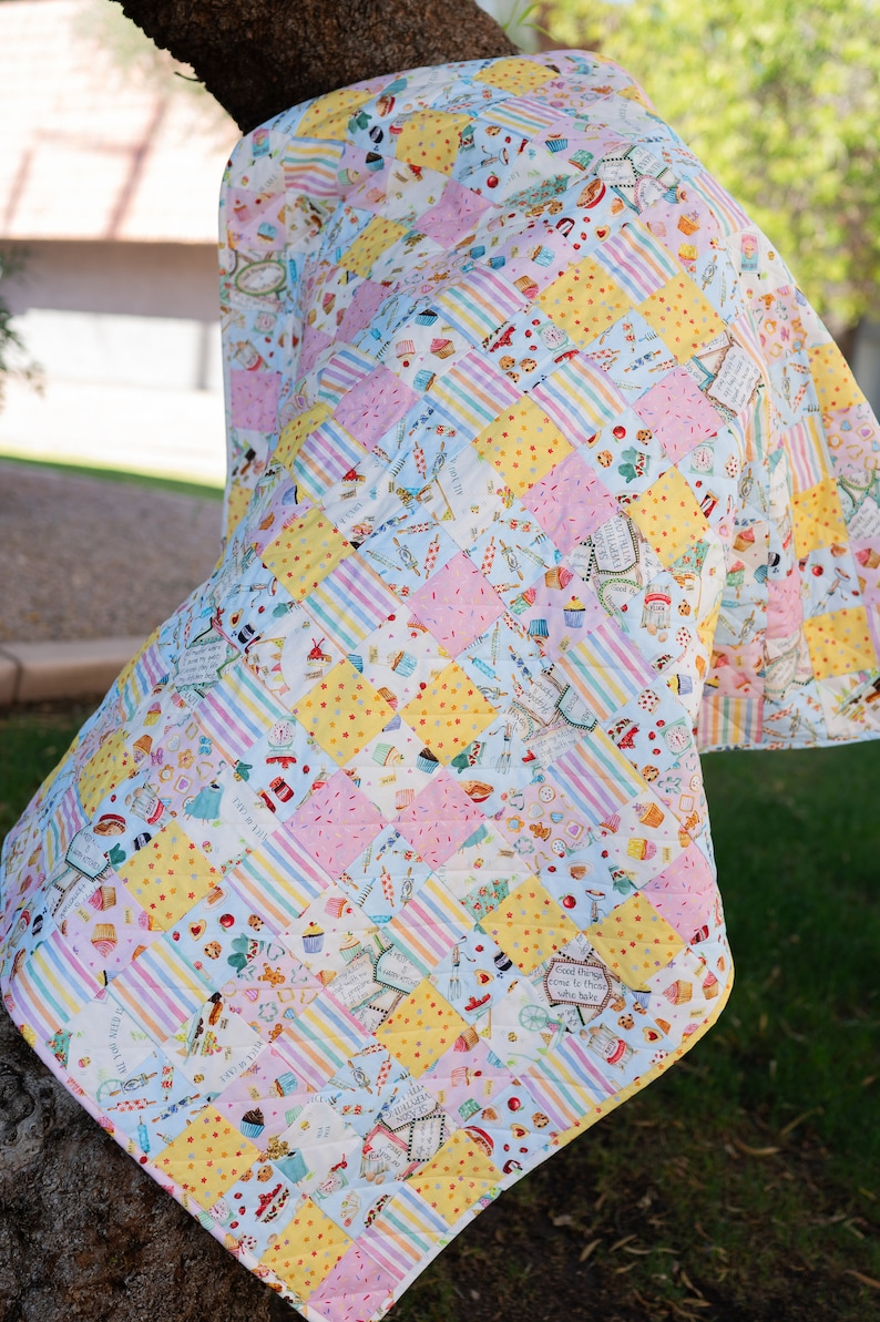 May include: A patchwork quilt with a light blue, yellow, and pink color scheme. The quilt is made of various fabrics with different patterns, including stripes, polka dots, and floral designs. The quilt is hanging on a tree branch.