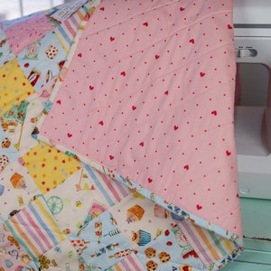 May include: A pink and white patchwork quilt with a variety of colorful patterns, including stripes, polka dots, and floral designs. The quilt is made of cotton fabric and has a quilted texture. The quilt is folded in half and is lying on a light blue surface.