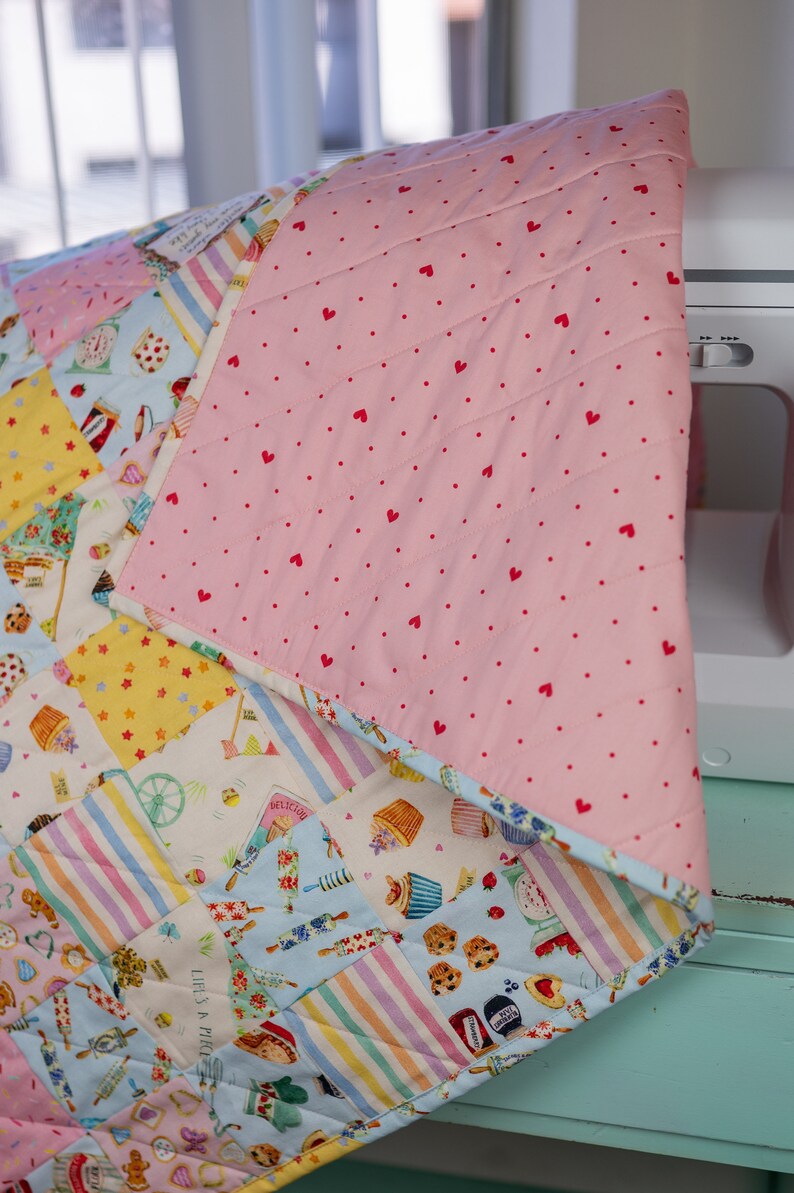 May include: A pink and white patchwork quilt with a variety of colorful patterns, including stripes, polka dots, and floral designs. The quilt is made of cotton fabric and has a quilted texture. The quilt is folded in half and is lying on a light blue surface.