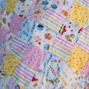 May include: A patchwork quilt with a variety of colorful fabrics featuring a whimsical baking theme. The quilt is made up of squares with different patterns, including cupcakes, sprinkles, stars, and stripes.