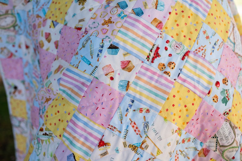 May include: A patchwork quilt with a variety of colorful fabrics featuring a whimsical baking theme. The quilt is made up of squares with different patterns, including cupcakes, sprinkles, stars, and stripes.