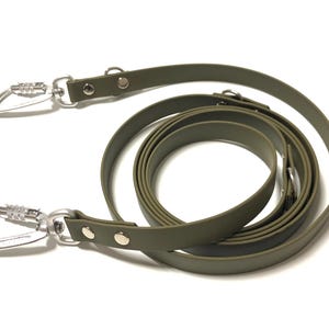 Op de afbeelding: Een olijfgroene hondenriem gemaakt van een glad, flexibel materiaal. De riem is voorzien van twee zilverkleurige metalen karabijnhaken aan beide uiteinden, vastgezet met zilveren klinknagels. De riem is in een cirkel opgerold met een matte afwerking.