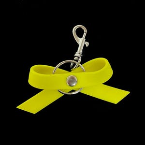 Könnte beinhalten: Ein hellgelber schlüsselanhänger in Schleifenform mit einem silberfarbenen Karabiner und Schlüsselring. Das Band ist mit einem silbernen Knopf befestigt. Der Hintergrund ist schwarz.