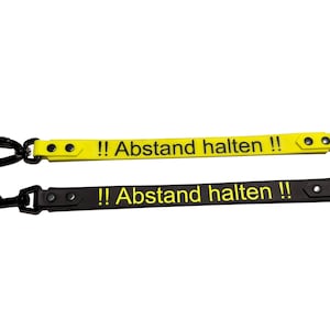 May include: Two dog leashes with the text "!! Abstand halten !!". One leash is yellow with a black carabiner and the other is black with a black clasp. Both leashes have a metal ring.