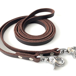 May include: A dark brown leather leash featuring two silver-toned metal swivel snap hooks at the ends. The leash is coiled into a circular shape, showing a smooth top surface and a textured underside, with metal rivets securing the hardware in place.