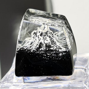 May include: A clear, rectangular resin keycap featuring a mountain scene. The bottom half is black, and the top half has a white mountain range. The keycap is set on a clear, faceted base.