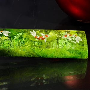Peut inclure: Objet rectangulaire en résine verte translucide avec un motif de poissons koï. Les koïs sont blancs et orange, sur un fond vert herbeux. L'objet est posé sur une surface noire réfléchissante.