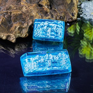 May include: Three blue rectangular resin pieces with white designs inside. The pieces are on a dark blue surface with a reflection of the pieces.