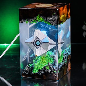 May include: A rectangular resin lamp featuring a white geometric drone-like figure suspended inside. The clear resin block contains dark wood accents and bright green moss-like textures. A white power cord extends from the side of the lamp.