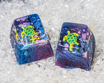 Pode incluir: Duas teclas coloridas com um design de tartaruga marinha. As teclas são feitas de resina e têm um padrão de redemoinho de azul, roxo e rosa. As tartarugas são verdes e amarelas e são pintadas em um estilo realista.
