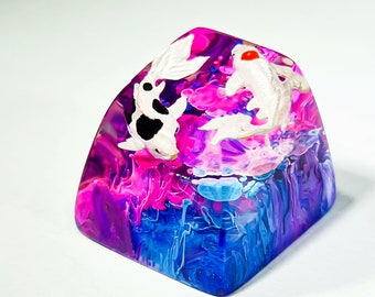 Pode incluir: Um keycap de resina com um design abstrato rosa, azul e branco com dois peixes koi brancos com marcas pretas.