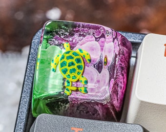 May include: A green and yellow turtle keycap with a pink and purple resin base. The keycap is on a black keyboard with white keys labeled "Tab", "Q", "1!" and "2".