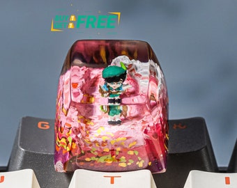 May include: A pink and gold resin keycap with a miniature figure of a person wearing a green hat and a pink dress. The keycap is sitting on a white keyboard with black keys.