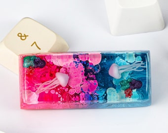 May include: A colorful resin keycap with a pink, blue, and white abstract design featuring two white jellyfish. The keycap is designed to fit a standard mechanical keyboard.