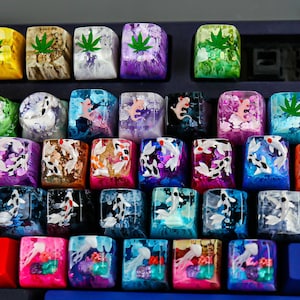 May include: A collection of artisan resin keycaps arranged on a mechanical keyboard. The keycaps feature embedded designs including green cannabis leaves, koi fish in various colours, axolotls, and jellyfish, set within translucent, multi-coloured bases.