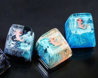 Pode incluir: Quatro teclas de resina com um design de axolotl rosa. Cada tecla tem um esquema de cores diferente: preto e azul, azul e branco, laranja e azul, e branco e preto.