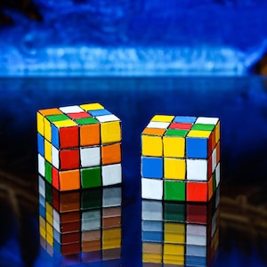 May include: Two colorful Rubik's Cubes with a variety of colored squares, including red, orange, yellow, green, blue, and white. The cubes are set against a reflective surface with a blue and brown background.