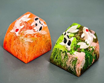 May include: Two resin keycaps with a koi fish design. One keycap is orange with white and black koi fish, the other is green with white and black koi fish.