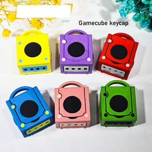 May include: A collection of six colorful Gamecube keycaps in yellow, purple, red, blue, pink, and green. Each keycap has a circular black center, a handle, and a rectangular base with three small holes. The text "Gamecube keycap" is visible.
