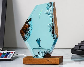 May include: A blue resin sculpture with a wooden base. The resin contains a depiction of a dolphin and a fish swimming in a blue ocean. The sculpture is lit from within, creating a warm glow.