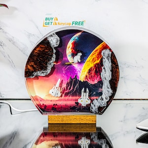 May include: A round resin lamp featuring a colourful, abstract space scene. The scene includes a planet, stars, and a spaceship. The lamp has a wooden base and is illuminated from within.