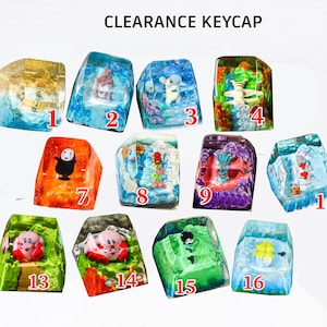 Puede incluir: Una colección de dieciséis teclas de resina transparente, cada una con una figura en miniatura y elementos de paisaje coloridos. El texto CLEARANCE KEYCAP está impreso en la parte superior. Cada tecla está numerada en color rojo.
