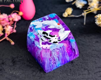 Pode incluir: Um keycap personalizado com um design abstrato roxo, azul e branco com dois peixes koi brancos com marcas pretas. O keycap é quadrado com bordas arredondadas.