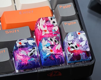 Pode incluir: Quatro teclas personalizadas com um design de peixe koi. As teclas são feitas de resina transparente com um redemoinho rosa, azul e roxo. Os peixes koi são pintados de branco e preto com detalhes vermelhos. As teclas estão em um teclado preto.