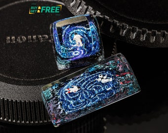 May include: Two small, square, resin keycaps with a blue, white, and black galaxy-like design. The keycaps are on a black surface.