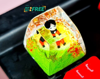 May include: A clear resin keycap with a green, orange, and yellow landscape design. A small cartoon figure is standing in the landscape. The keycap is on a black keyboard with red keys.