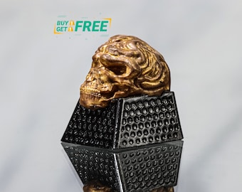 Puede incluir: Un keycap de calavera dorado con detalles intrincados se asienta sobre una base de keycap negra y gris. El keycap es un elemento decorativo para un teclado mecánico.