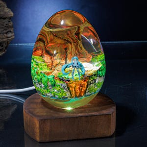 Puede incluir: Una l&aacute;mpara de vidrio transparente en forma de huevo con una base de madera. La l&aacute;mpara contiene una figura en miniatura de un personaje de anime de pelo azul de pie en un paisaje verde y marr&oacute;n. La l&aacute;mpara est&aacute; iluminada desde abajo, proyectando un brillo c&aacute;lido.
