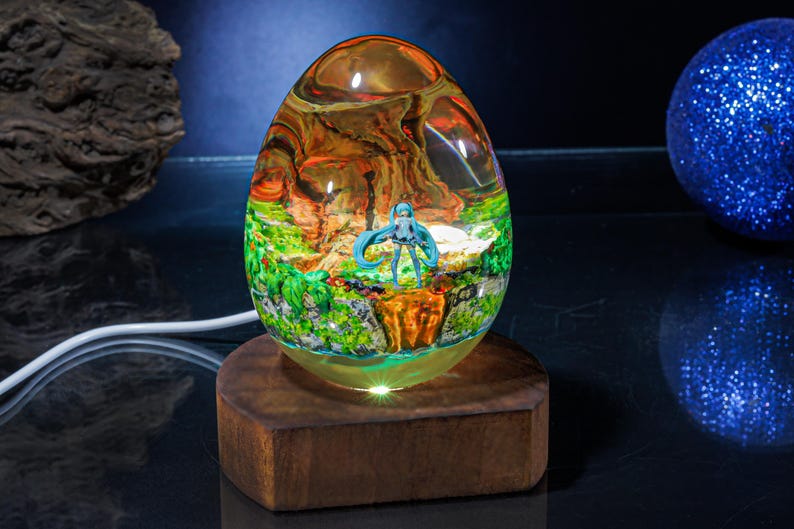 Puede incluir: Una l&aacute;mpara de vidrio transparente en forma de huevo con una base de madera. La l&aacute;mpara contiene una figura en miniatura de un personaje de anime de pelo azul de pie en un paisaje verde y marr&oacute;n. La l&aacute;mpara est&aacute; iluminada desde abajo, proyectando un brillo c&aacute;lido.