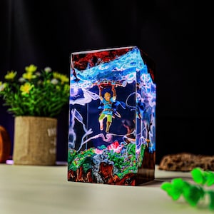 May include: A resin cube featuring a miniature figure of a video game character suspended within. The cube has a layered design with red, blue, and green elements, creating a vibrant, artistic display. The figure is holding a brown object.