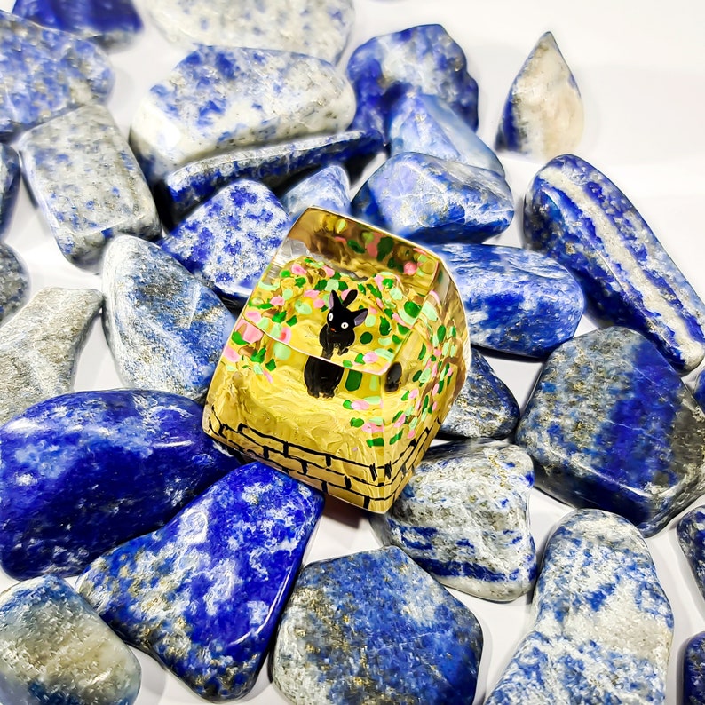 K&ouml;nnte beinhalten: Ein Harz-Tastenkappen mit einem schwarzen Katzen-Design, umgeben von blauen und wei&szlig;en Steinen. Die Tastenkappe ist gelb mit einem Ziegelmuster und einem Blumendesign.