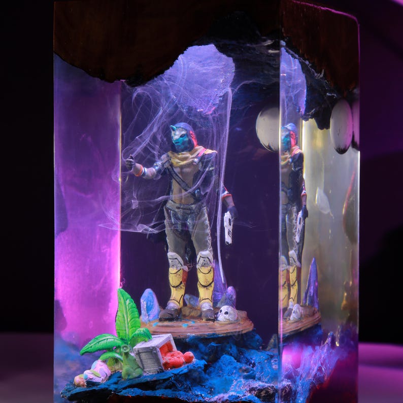 May include: A rectangular resin block containing a miniature figure of a character with a blue mask, yellow leg armor, and a white pistol. The scene includes a small green plant, a white crate, and a skull, all illuminated by purple light.