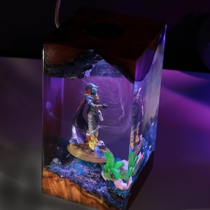 May include: A rectangular resin lamp featuring a dark wood top and base with a clear center section. Inside the resin, a miniature figure stands on a circular platform surrounded by small green plants and blue accents, illuminated by a purple light.