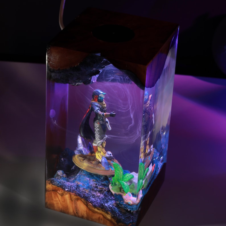 May include: A rectangular resin lamp featuring a dark wood top and base with a clear center section. Inside the resin, a miniature figure stands on a circular platform surrounded by small green plants and blue accents, illuminated by a purple light.