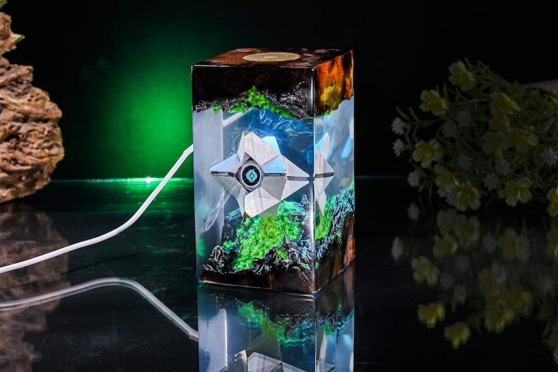 May include: A rectangular resin lamp featuring a white geometric drone-like figure suspended inside. The clear resin block contains dark wood accents and bright green moss-like textures. A white power cord extends from the side of the lamp.