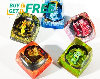 May include: Five colorful resin keycaps with different cartoon characters inside. The keycaps are square and have a glossy finish. The characters are a green monster, a red monster, a blue monster, a yellow monster, and a purple monster.