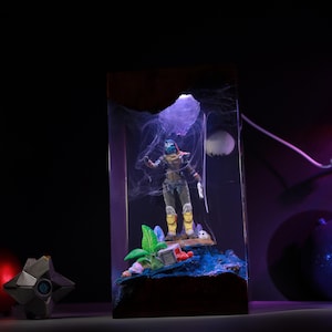 May include: A rectangular resin lamp featuring a miniature Destiny character figure standing on a textured blue base with green foliage. The scene is illuminated by a purple light, creating a smoky effect around the figure, with a small white sphere resembling a moon in the background.