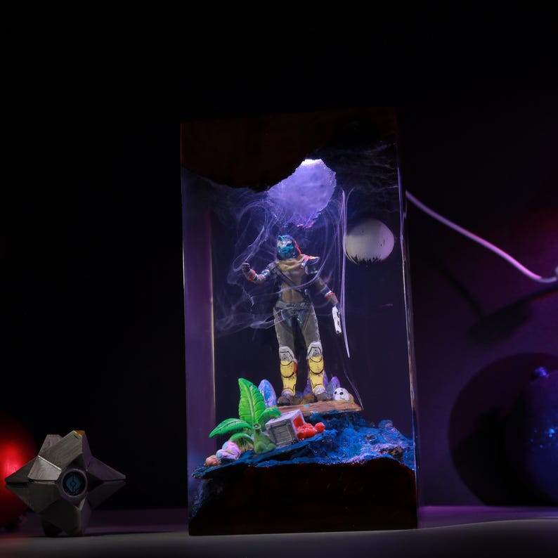 May include: A rectangular resin lamp featuring a miniature Destiny character figure standing on a textured blue base with green foliage. The scene is illuminated by a purple light, creating a smoky effect around the figure, with a small white sphere resembling a moon in the background.