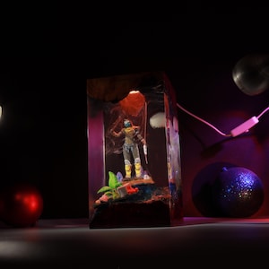May include: A rectangular resin lamp containing a miniature figure of a character in yellow and grey armor standing on a rocky base with green foliage. The interior is illuminated with a warm red glow, creating a smoky effect around the figure.
