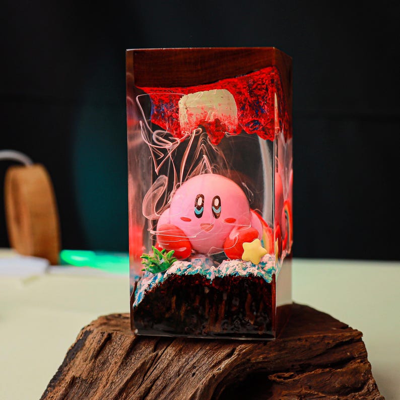 May include: A pink Kirby figurine inside a clear resin block with a wooden base. The block features a red and white design at the top, with a star and green plant accents near the figurine. The background is dark.